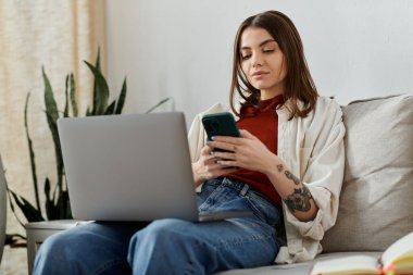 Dizüstü bilgisayarı ve akıllı telefonuyla koltukta oturan genç bir kadın işine odaklanıyor..