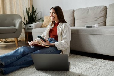 Günlük giysiler içindeki genç bir kadın oturma odasında yerde oturuyor, dizüstü bilgisayarı ve telefonu üzerinde çalışıyor..