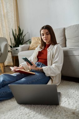 Günlük kıyafetli genç bir kadın dizüstü bilgisayarı ve elinde telefonuyla yerde oturuyor ve evden çalışıyor..