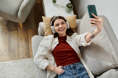 Günlük kıyafetli genç bir kadın koltukta dinleniyor, kulaklıkları takılı, telefonunda selfie çekiyor..