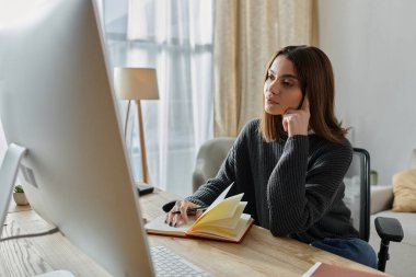 Gri kazaklı genç bir kadın çalışma masasında oturuyor, bir dizüstü bilgisayar üzerinde çalışıyor ve bir deftere yazı yazıyor..