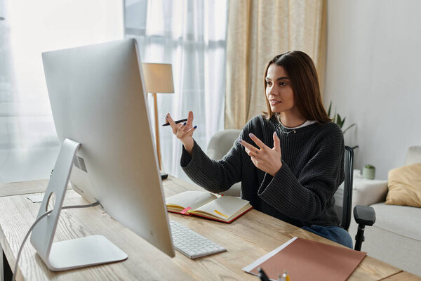 Молодая женщина в сером свитере создает увлекательный цифровой контент из своего домашнего офиса, страстно разговаривая на экране своего компьютера.