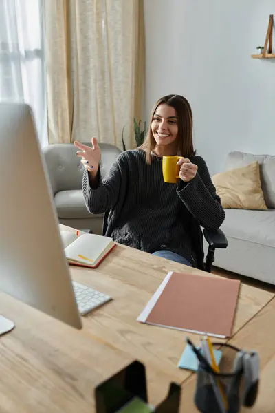 stock image A young woman in a gray sweater sits at her desk at home, holding a cup of coffee and working on her computer.