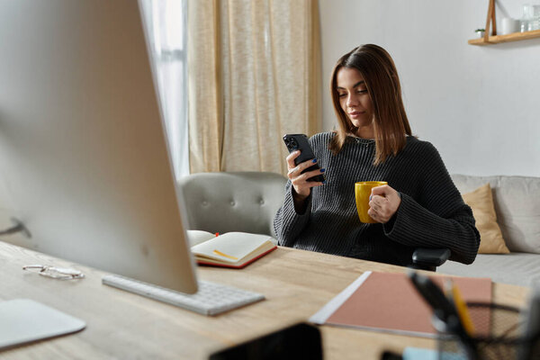 Женщина работает из дома, приклеена к телефону, потягивает кофе, вероятно, управляет цифровым маркетингом.