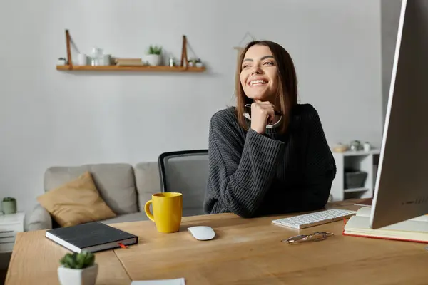 stock image A young woman in a grey sweater sits at her desk in a home office, smiling and looking thoughtfully ahead.