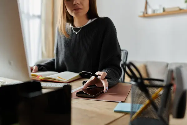 stock image A young woman in a grey sweater sits at a desk working on her laptop and phone, focusing on her work.