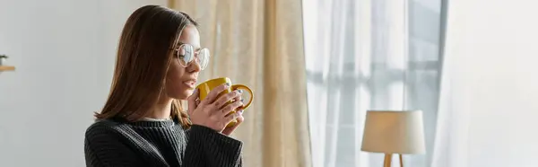 stock image A young woman wearing a grey sweater and glasses enjoys a cup of coffee while looking out the window.