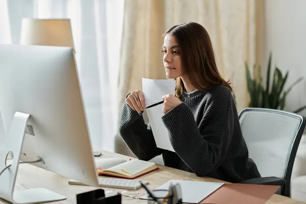 stock image A young woman in a grey sweater works on her laptop at a desk, reviewing ideas for her digital marketing content.