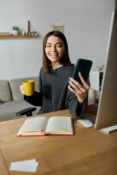 stock image A young woman sits at a desk in a home office, smiling as she records a video on her phone.