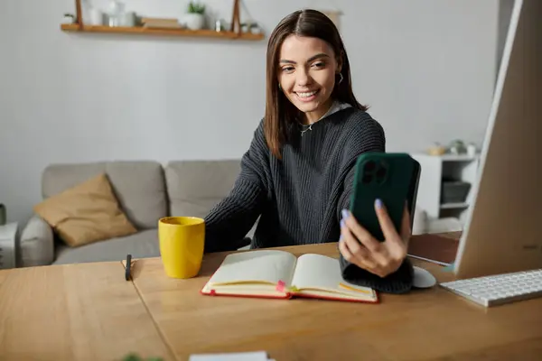 stock image A young woman works remotely from home, smiling as she uses her phone to create content.