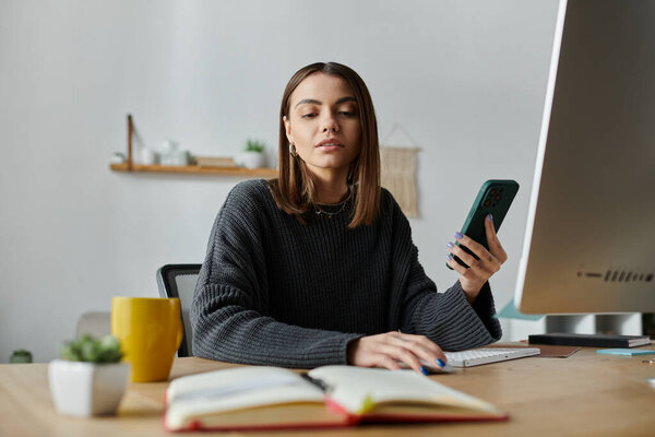 Молодая женщина в сером свитере сидит за столом в своем домашнем офисе, работая над ноутбуком, держа в руке телефон..