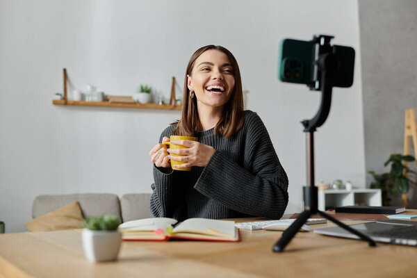 Молодая женщина сидит за столом в своем доме, записывая контент на телефон, счастливо улыбаясь и держа чашку.