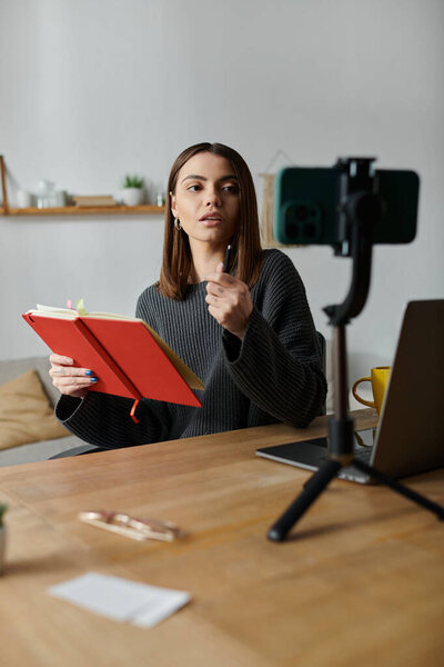 Молодая женщина в сером свитере сидит за своим столом, записывая видео на свой телефон, держа в руках красный блокнот и глядя в камеру.