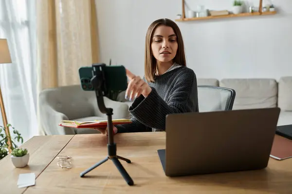 stock image A young woman works remotely, filming a video on her phone while sitting at her desk with a laptop.