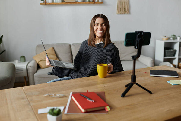 Молодая женщина сидит за столом с ноутбуком, чашкой кофе и телефоном на штативе, создавая контент для своей аудитории.