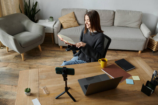 Молодая женщина сидит за столом, работает над ноутбуком, записывает видео и делает заметки.