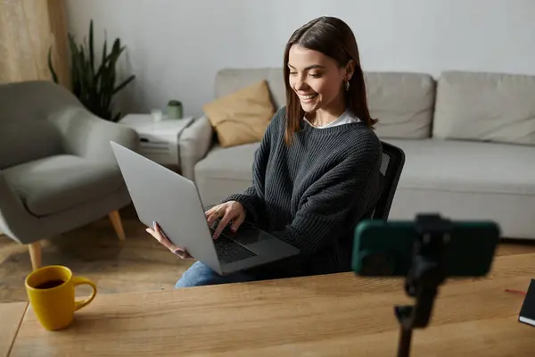 stock image A young woman in a grey sweater works on her laptop at a desk while filming a video.