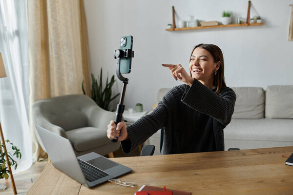 A young woman films herself with a phone on a selfie stick while sitting at her home office desk.