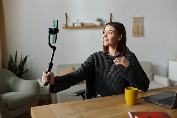 stock image A young woman wearing a gray sweater smiles as she films content with a smartphone attached to a selfie stick.