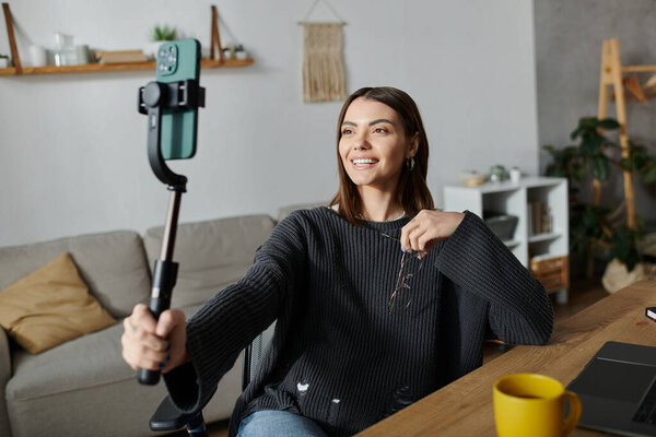 Молодая женщина, одетая в серый свитер, сидит за столом в своем доме, используя селфи-палку, чтобы снять контент на телефон..