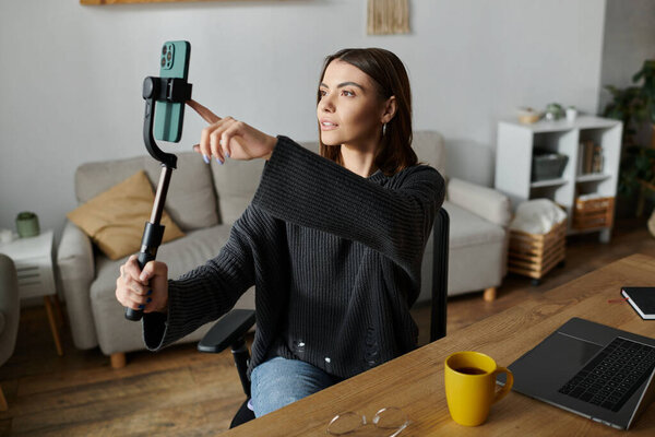 Молодая женщина в сером свитере, использующая телефон, прикрепленный к селфи-палочке.