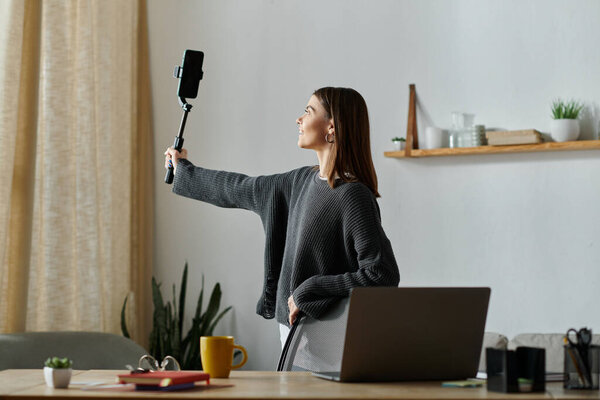 Молодая женщина в сером свитере, довольствующаяся смартфоном и селфи-палкой, стоя за своим рабочим столом.