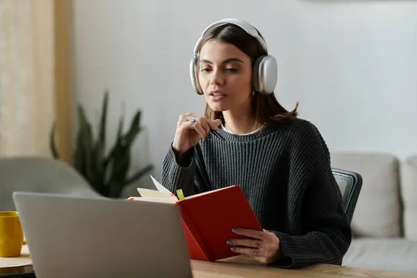 stock image A young woman in a grey sweater is working from home, sitting at a desk with a laptop and a notebook.