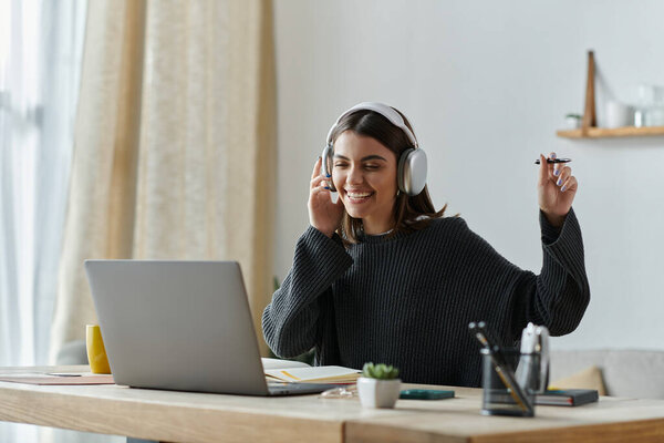 A young woman smiles while working on her laptop at her home office, listening to music and tapping a pen to the beat.