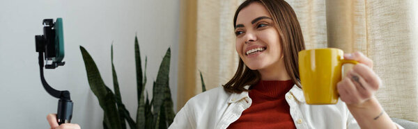 Молодая женщина в повседневной одежде держит стабилизатор смартфона и записывает видеоблог, наслаждаясь чашкой кофе.