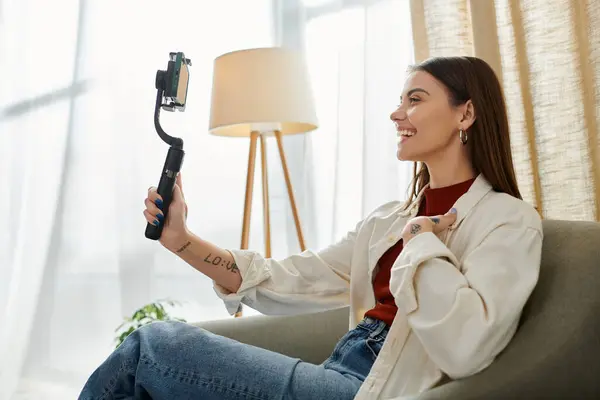 stock image A young woman, casually dressed, sits on a couch and vlogs while holding a smartphone stabilizer.
