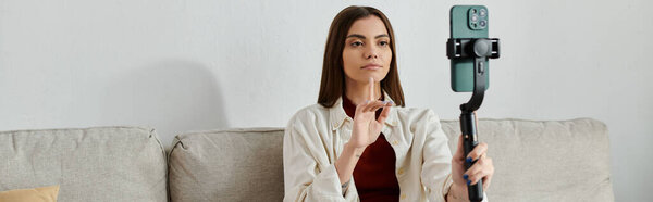 Молодая женщина сидит на диване, держит стабилизатор смартфона и снимает видеоблог.