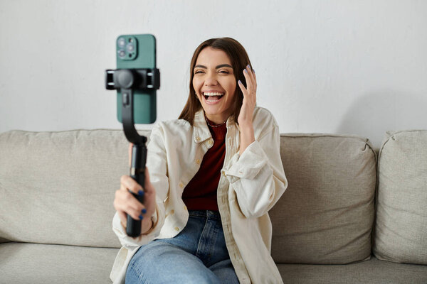 A young woman with a bright smile sits on a couch, filming a vlog with her smartphone and stabilizer.