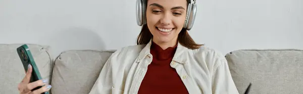 stock image A young woman in casual attire sits on a sofa, listening to music through wireless headphones and holding a phone.