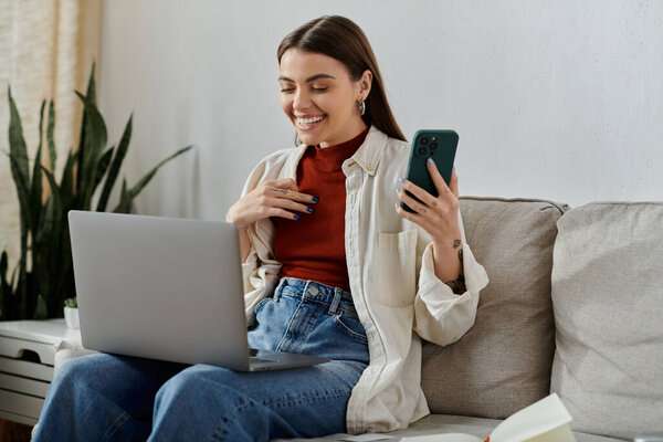 Молодая женщина в повседневной одежде сидит на диване, работает на ноутбуке и проверяет телефон с улыбкой.