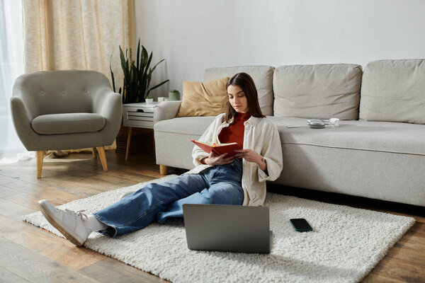 A young woman wearing casual clothes sits on the floor in a living room, working on her laptop and reading a notebook.