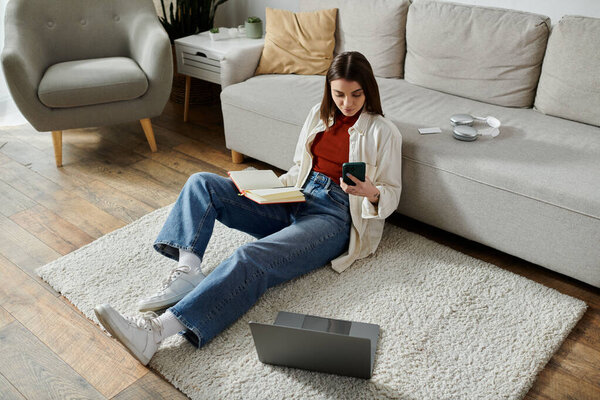 A woman in jeans and a white shirt sits on a rug in her living room, working on her laptop and scrolling through her phone.