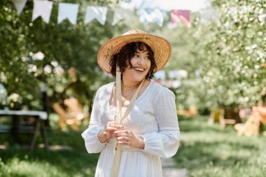 A young woman with dark hair and a straw hat smiles brightly in a sunny summer garden, dressed in a white blouse. clipart