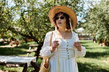 A brunette woman in a white dress and a straw hat, enjoying the sunshine in a green summer garden. clipart