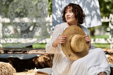 Beyaz elbiseli ve hasır şapkalı genç bir kadın bir yaz bahçesinde oturuyor, düşüncelere dalmış..