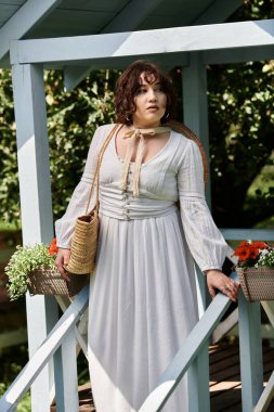 Beyaz elbiseli ve hasır şapkalı genç bir kadın bahçe yolunda yürüyor, bakışları mesafeye sabitlenmiş..