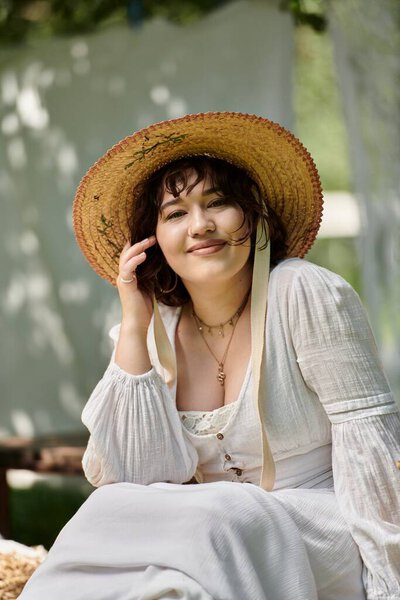 A brunette woman smiles sweetly while wearing a straw hat and a white blouse in a garden setting.