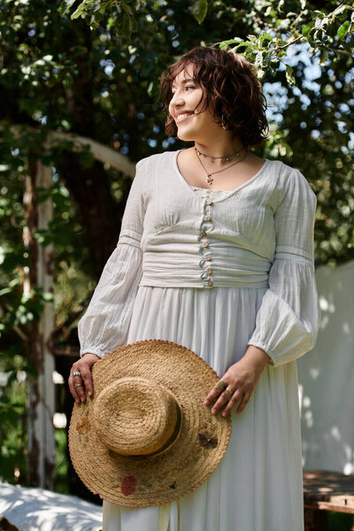 A young woman in a white dress and straw hat stands in a summer garden, her smile bright against the greenery.