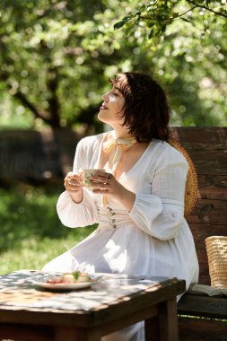 Beyaz elbiseli genç bir kadın bereketli yeşilliklerle çevrili bir yaz bahçesinde hafif bir brunch 'ın tadını çıkarıyor..