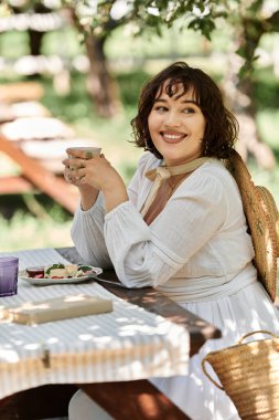 Beyaz elbiseli genç bir kadın yemyeşil bir bahçede yaz brunch 'ının tadını çıkarıyor..