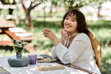 Beyaz elbiseli genç bir kadın bereketli bir yaz bahçesinde rahatlatıcı bir brunch 'ın tadını çıkarıyor..
