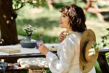 Beyaz elbiseli genç bir kadın açık havada yemek yiyor, yanında bir kitap ve hasır şapkayla bir masada oturuyor..