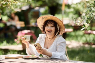 A young woman in a white dress enjoys a summer garden brunch, smiling while holding a cup of tea and her phone. clipart