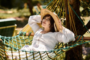 Beyaz elbiseli ve hasır şapkalı genç bir kadın yaz gökyüzünün altında bir hamakta dinleniyor..