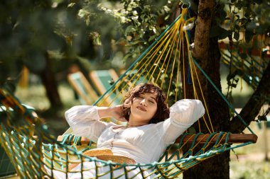 Beyaz elbiseli ve hasır şapkalı genç bir kadın hamakta dinleniyor, yemyeşil bir bahçede yaz güneşinin tadını çıkarıyor..