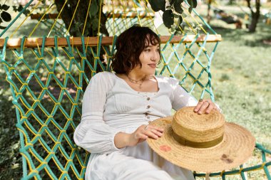 A young woman in a white dress and straw hat relaxes in a hammock under the shade of a tree. clipart